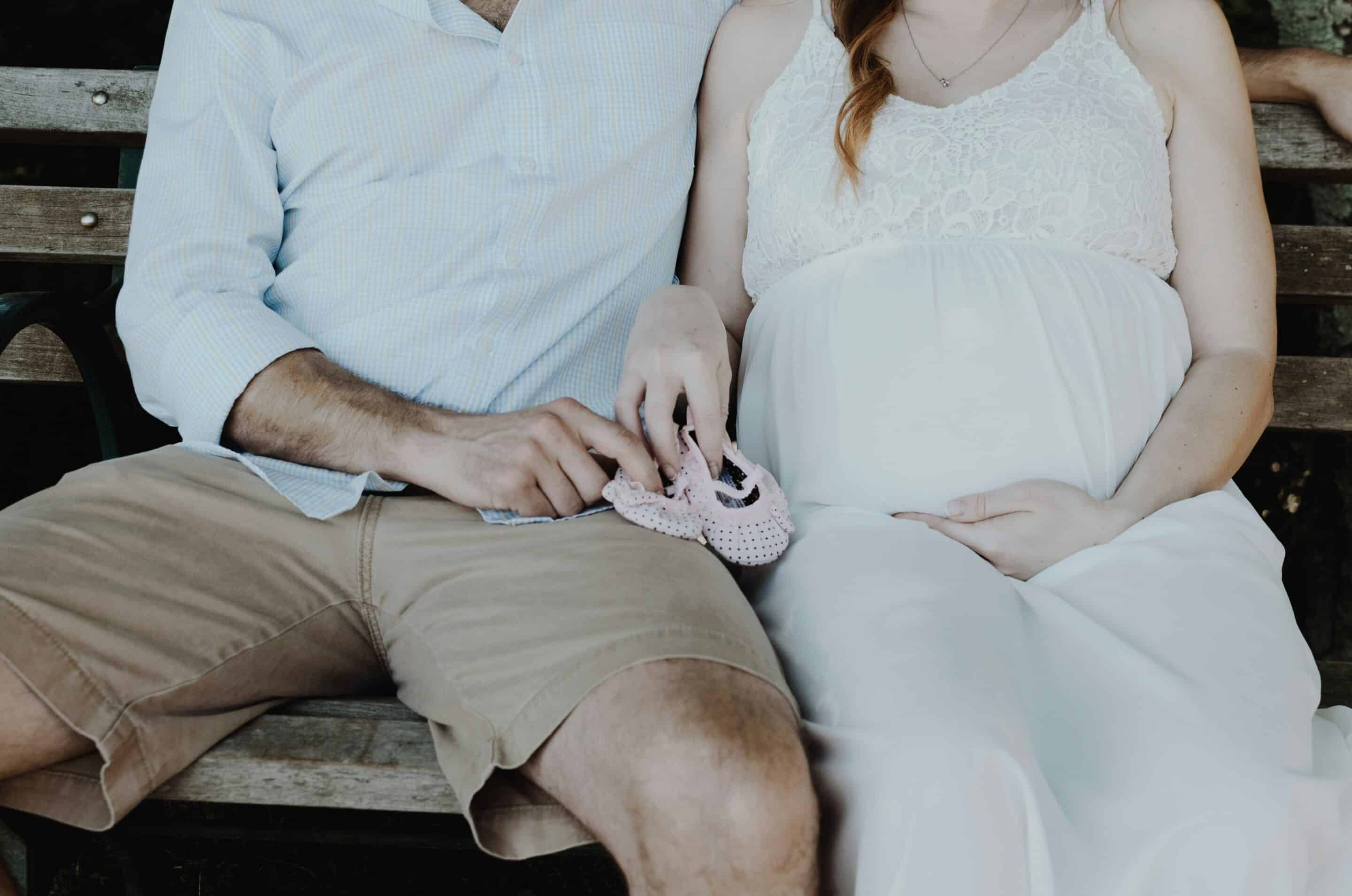 Una donna in gravidanza seduta accanto al partner con un paio di scarpine da neonato, immagine legata alla sessualità in gravidanza.